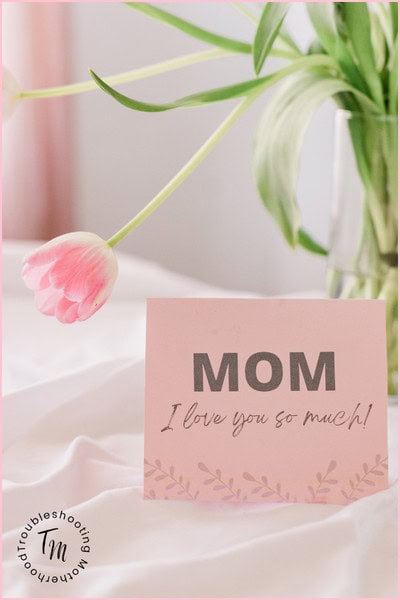 A pink card that reads "MOM I love you so much!" stands on a white surface next to a vase of pink tulips. mother's day gifts for mom