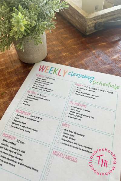 A printed weekly cleaning schedule is displayed on a wooden surface, outlining daily and weekly tasks for stay-at-home moms, including laundry, kitchen cleaning, and organization tips.