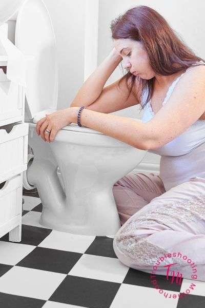A pregnant woman sits on the floor beside a toilet, appearing distressed, which may suggest a moment of experiencing morning sickness.