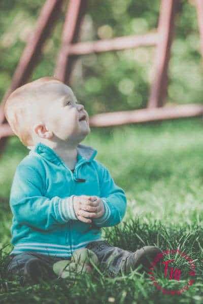 A young boy in a blue sweater sits on the grass, looking upwards with a joyful expression, framed by a blurred swing set in the background.