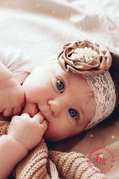 A close-up photograph of a baby girl with blue eyes, wearing a lace headband adorned with a floral design and pearls, while grasping a piece of fabric.