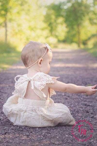 A baby girl wearing a light-colored dress sits on a gravel path surrounded by greenery, embodying the essence of cute names for girls and kid name ideas.