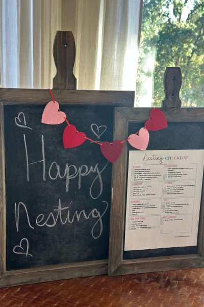 Two rustic wooden chalkboards decorated with a red and pink heart garland, one reading “Happy Nesting” and the other displaying a printed nesting party checklist. A cozy, sunlit background sets the scene for a welcoming nesting baby shower or nesting party.