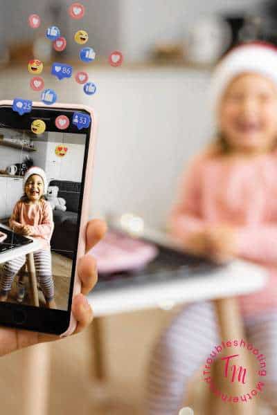 Parent taking a photo of a smiling child with social media reaction icons floating up, highlighting online privacy and social media boundaries for kids.
