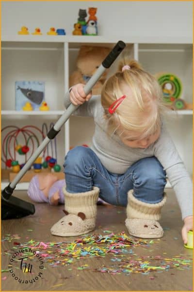Toddler helping clean by sweeping confetti off the floor with a toy broom in a playroom while learning to help with chores.